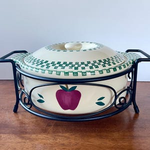 May include: A white ceramic casserole dish with a green and white checkered pattern and an apple design. The dish is on a black metal stand with a decorative scroll design.