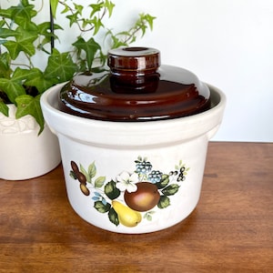 May include: A white ceramic crock with a brown lid and a floral design featuring apples, pears, and berries. The crock has a speckled glaze and a brown rim.