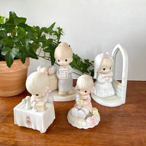 Puede incluir: Conjunto de cuatro figuritas de porcelana de ni&ntilde;os vestidos de blanco y rosa. Las figuritas sostienen libros, bloques y un peque&ntilde;o p&aacute;jaro blanco. Las figuritas est&aacute;n sobre una base blanca con un dise&ntilde;o floral rosa y blanco.