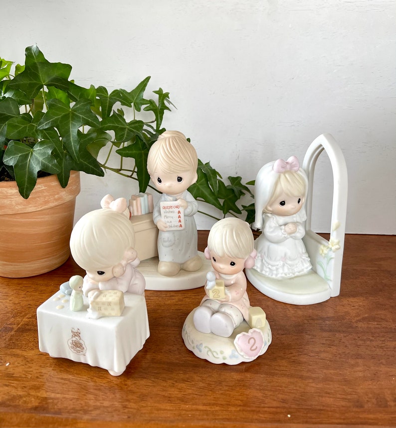 Puede incluir: Conjunto de cuatro figuritas de porcelana de ni&ntilde;os vestidos de blanco y rosa. Las figuritas sostienen libros, bloques y un peque&ntilde;o p&aacute;jaro blanco. Las figuritas est&aacute;n sobre una base blanca con un dise&ntilde;o floral rosa y blanco.