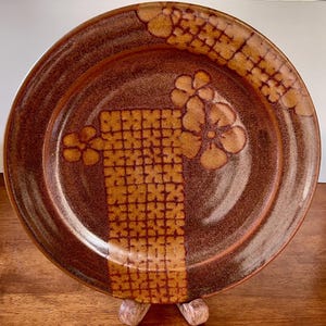 May include: A brown ceramic plate with a floral and grid pattern. The plate has a dark brown rim and a lighter brown center. The floral design is in a lighter shade of brown.