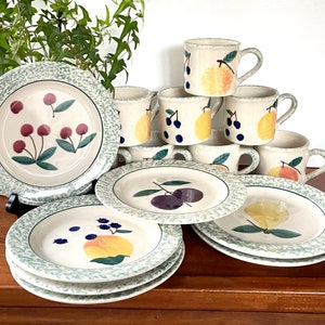 May include: A set of green and white ceramic plates and mugs with fruit designs. The plates have a green rim and a white center with a design of cherries, plums, or peaches. The mugs have a similar design and are stacked on top of each other.