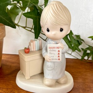 Puede incluir: Una figurita de porcelana de un ni&ntilde;o sosteniendo una tarjeta de calificaciones con todas las A. El ni&ntilde;o lleva un vestido azul y blanco y tiene el pelo rubio. La tarjeta de calificaciones dice "Report Card" y enumera las materias "Kindness", "Mercy", "Love" y "Truthfulness". La figurita est&aacute; de pie sobre una peque&ntilde;a base con forma de coraz&oacute;n.