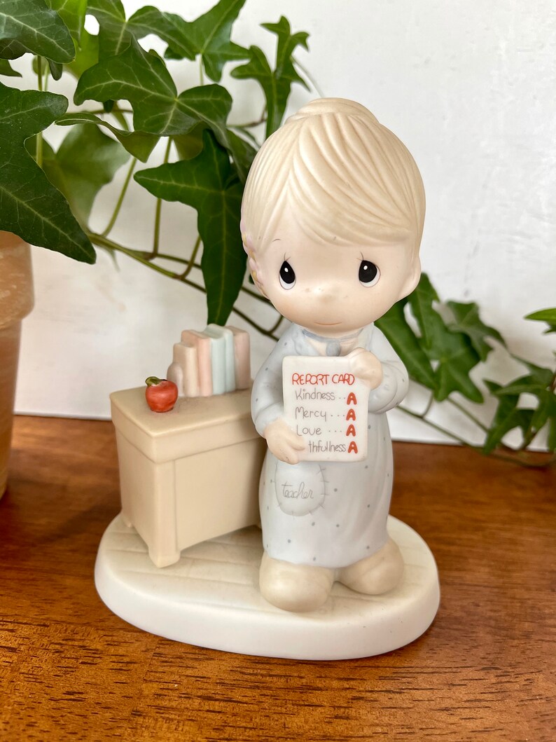 Puede incluir: Una figurita de porcelana de un ni&ntilde;o sosteniendo una tarjeta de calificaciones con todas las A. El ni&ntilde;o lleva un vestido azul y blanco y tiene el pelo rubio. La tarjeta de calificaciones dice "Report Card" y enumera las materias "Kindness", "Mercy", "Love" y "Truthfulness". La figurita est&aacute; de pie sobre una peque&ntilde;a base con forma de coraz&oacute;n.