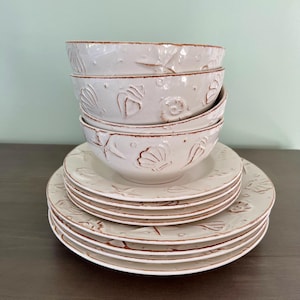 May include: A stack of off-white ceramic bowls and plates with embossed seashell designs. The bowls and plates have a rustic, textured finish with brown trim. The set includes four bowls and six plates, perfect for a coastal-themed dining experience.