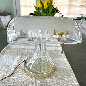 May include: A clear glass table lamp with a frosted shade and a gold-colored base. The lamp has a decorative floral pattern on the base and a pull chain. Yellow tulips are in a vase behind the lamp.