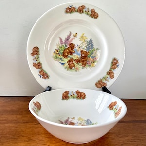May include: A white ceramic plate and bowl set featuring a charming bear and bunny design. The plate has a border of bears and a central scene with bears, a bunny, and flowers. The bowl mirrors the design, creating a cohesive children's tableware set.