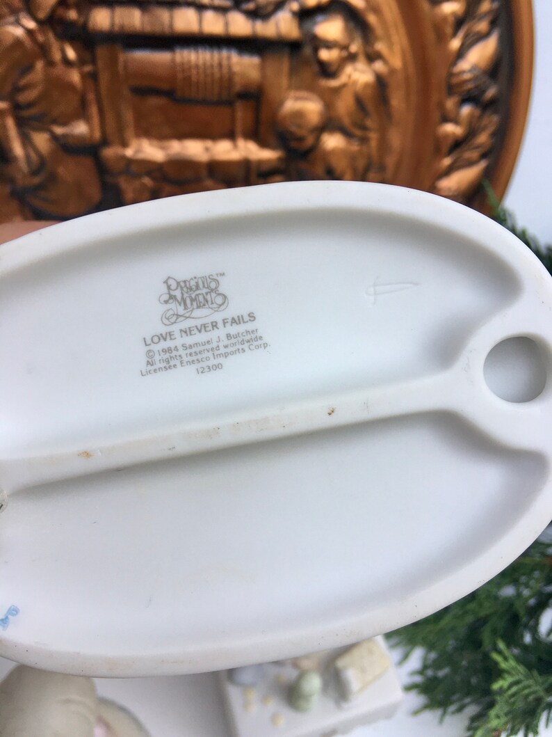 Puede incluir: Una figurita de porcelana blanca con un borde dorado. La figurita representa a una pareja tom&aacute;ndose de la mano y mir&aacute;ndose. La base de la figurita tiene las palabras "Precious Moments" y "Love Never Fails" impresas. La figurita est&aacute; hecha por Enesco Imports Corp. y tiene un n&uacute;mero de licencia de 12300.