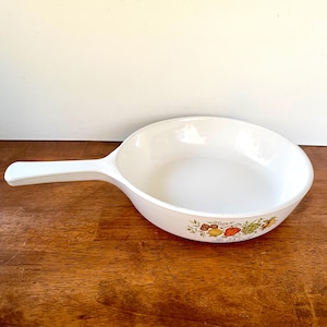 May include: White ceramic skillet with a long handle. The skillet has a decorative band of colorful fruits and vegetables around the rim. The skillet is on a wooden surface.