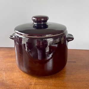May include: A brown ceramic crock pot with a lid. The crock pot has a smooth, glossy finish and a slightly rounded shape. The lid has a knob on top.