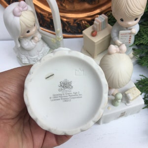 Puede incluir: Un conjunto de figuras de porcelana blanca con tres personajes de Precious Moments. Los personajes son una ni&ntilde;a con un lazo rosa en el pelo, una maestra con un libro y una ni&ntilde;a que sostiene un libro. El conjunto forma parte de la colecci&oacute;n Precious Moments y se titula "Growing In Grace, Age 2".