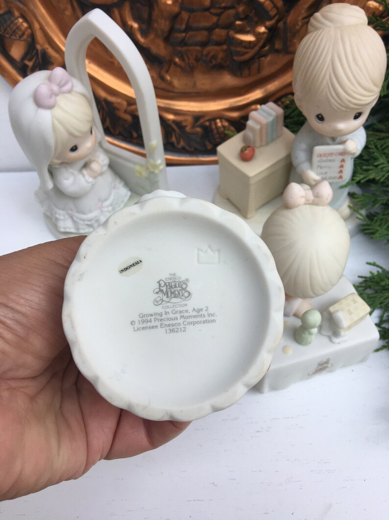 Puede incluir: Un conjunto de figuras de porcelana blanca con tres personajes de Precious Moments. Los personajes son una ni&ntilde;a con un lazo rosa en el pelo, una maestra con un libro y una ni&ntilde;a que sostiene un libro. El conjunto forma parte de la colecci&oacute;n Precious Moments y se titula "Growing In Grace, Age 2".
