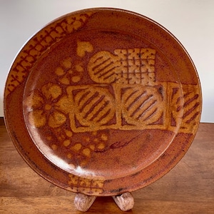 May include: A brown ceramic plate with a raised geometric design. The plate has a textured surface and a slightly raised rim.