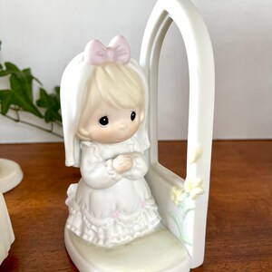 Puede incluir: Una figurita de porcelana blanca de una ni&ntilde;a arrodillada en oraci&oacute;n con las manos juntas. Lleva un vestido blanco con un lazo rosa y un velo blanco. La figurita est&aacute; colocada frente a un arco blanco con un dise&ntilde;o floral.