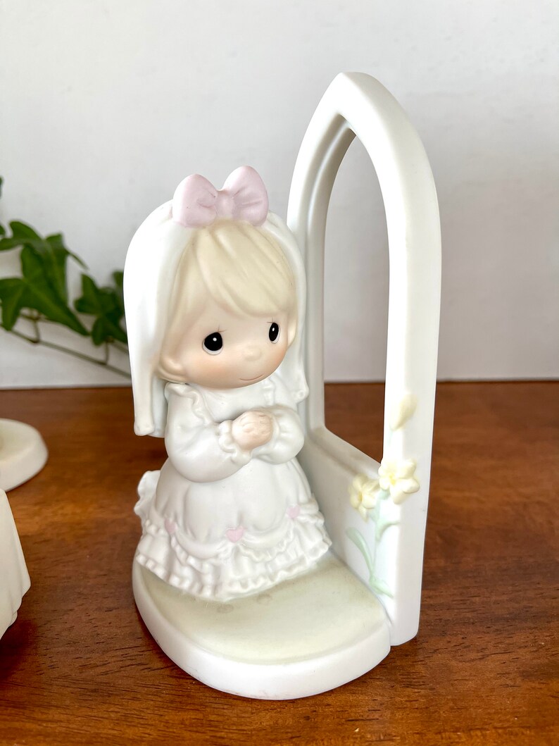 Puede incluir: Una figurita de porcelana blanca de una ni&ntilde;a arrodillada en oraci&oacute;n con las manos juntas. Lleva un vestido blanco con un lazo rosa y un velo blanco. La figurita est&aacute; colocada frente a un arco blanco con un dise&ntilde;o floral.