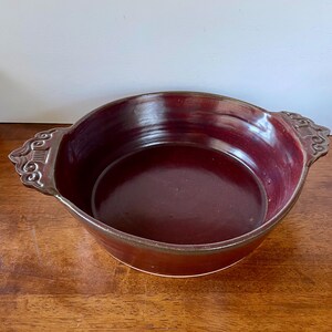 May include: A deep red ceramic baking dish with two decorative handles. The dish is glazed and has a slight sheen.