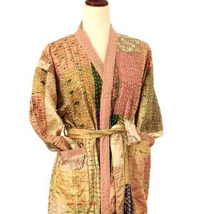 May include: A patchwork robe in shades of gold, pink, and green, with a matching belt. The robe features a quilted design and a light pink trim along the edges and belt. The robe is displayed on a mannequin.