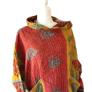 May include: A patchwork hooded tunic in shades of red, yellow, and teal. The tunic features a quilted design with floral and paisley patterns. It has two front pockets and a contrasting floral hem.