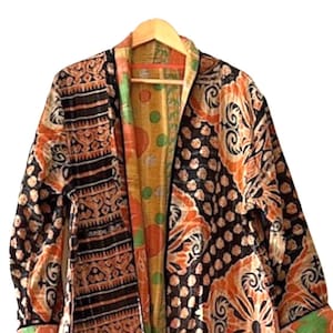 May include: A long, open-front robe with a vibrant, patterned design. The robe features a mix of black, orange, and cream colors with floral and geometric motifs. The interior lining is a contrasting yellow with polka dots and other patterns. The robe is hanging on a wooden hanger.