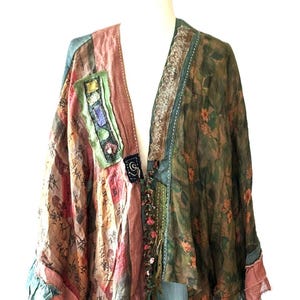 May include: A patchwork kimono-style jacket featuring a mix of fabrics. The left side displays red and tan panels with black calligraphy text, while the right side features a dark green floral print. It includes lace trim, decorative stitching, and frayed edges.