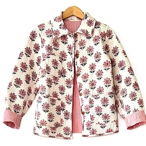 May include: A white jacket with a floral pattern in shades of pink and brown. The jacket has a collared neckline, button closures, and two front pockets. The cuffs and the inside lining are a solid pink color. The jacket is hanging on a wooden hanger.