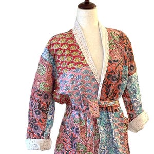 May include: A patchwork robe with a floral pattern in shades of red, pink, blue, and green. The robe has a white trim and a matching belt. The sleeves are three-quarter length, and the robe is displayed on a mannequin.