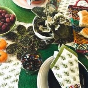May include: A table setting with green placemats, white plates, and green napkins. There is a small plant in a white pot, a bowl of grapes, and a glass of colorful beads on the table.