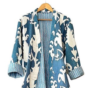 May include: A blue and white patterned kimono-style jacket with three-quarter sleeves. The jacket is made of a quilted fabric and has a striped lining. It hangs on a wooden hanger against a white background. The jacket has a tie closure.