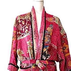 May include: A pink patchwork robe with a tie belt. The robe features a mix of floral and geometric patterns in shades of pink, yellow, and blue. Black trim outlines the edges and cuffs. The robe is displayed on a white background.
