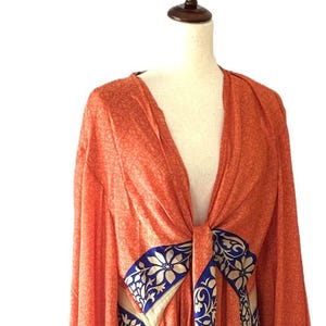 May include: A long-sleeved orange kimono featuring a subtle tonal pattern throughout. The garment includes contrasting panels with a blue and cream floral and paisley print along the front opening and hem, tied at the center with a fabric sash.