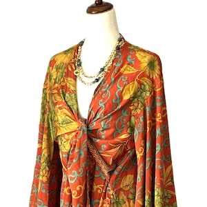 May include: A long-sleeved wrap top featuring a vibrant floral pattern in shades of orange, yellow, and green with turquoise scrollwork accents. The garment has a deep V-neckline and a front tie closure, displayed on a mannequin with a beaded necklace.