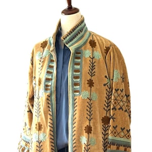 May include: A long, tan-colored coat featuring intricate embroidery in shades of light blue, dark brown, and cream. The garment has a high collar, long sleeves, and a vertical pattern of floral and geometric motifs running down the front panels.