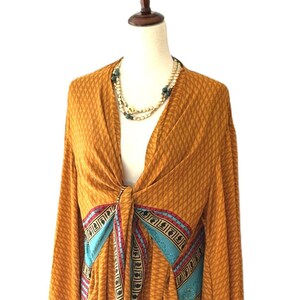 May include: A mustard yellow kimono-style wrap top featuring a repeating diamond pattern, long wide sleeves, and a front tie closure. The garment is accented with vertical panels of turquoise, red, and black geometric border prints along the front.