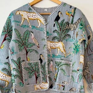 May include: A light gray quilted jacket featuring a vibrant jungle print with cheetahs, zebras, toucans, and palm trees. The jacket has a V-neckline, long sleeves, two front patch pockets, and is finished with a white and gray striped trim.