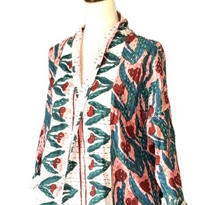 May include: A long-sleeved jacket featuring a repeating floral pattern in shades of dark red, teal, and light pink. The garment has a wide collar, an open front, and visible white running stitch details throughout the fabric construction.