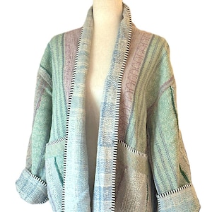 May include: A long-sleeved open-front jacket featuring a patchwork design in shades of light green, blue, and lavender. The garment includes two front pockets and is finished with a black and white striped trim along the collar and cuffs.