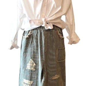 May include: White button-down shirt tied at the waist over a pair of distressed, striped, and patched denim trousers.