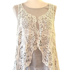 May include: A sleeveless, cream-colored lace top with a flowing, open front design. The intricate lace overlay sits atop a gray, layered garment, creating a delicate and airy aesthetic. The top is displayed on a mannequin.