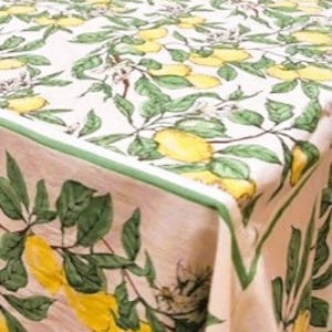 May include: A close-up of a tablecloth with a lemon and leaf pattern. The fabric is off-white with yellow lemons and green leaves. The border has a green trim. The tablecloth is on a table.