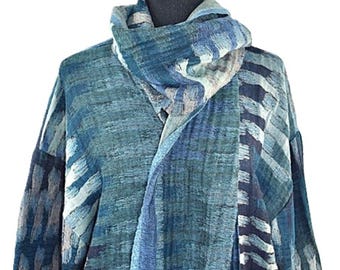 May include: A long-sleeved open-front cardigan featuring a textured, woven fabric in various shades of blue, teal, and grey. The garment includes an integrated scarf or shawl collar that drapes around the neck, displaying a consistent abstract pattern.