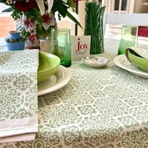 May include: A table setting featuring a light green and white patterned tablecloth, a folded napkin, a lime green bowl on a white plate, a green glass, a green pitcher, a small white dish, and a card with the text joy of cooking printed on it.