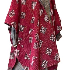 May include: A deep red hooded poncho featuring a repeating white diamond pattern and a visible blue and grey interior lining. The garment is worn over a grey collared shirt and dark pants, showcasing a textured fabric with visible stitching details.