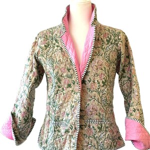 May include: A quilted jacket with a floral pattern in shades of green, pink, and beige. The jacket features a pink lining and cuffs, with black and white striped trim along the edges. The jacket is displayed on a mannequin.