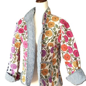 May include: A floral print jacket with a white background, featuring orange, pink, and purple flowers. The jacket has a striped blue and white lining and trim. The sleeves are three-quarter length, and the jacket has two pockets.