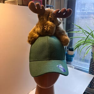 Könnte beinhalten: Eine einfarbige dunkelgrüne Baseballkappe mit einem aufgesetzten braunen Plüsch-Elch. Die Kappe hat eine strukturierte Krone und einen gebogenen Schirm mit einem kleinen rechteckigen Aufkleber auf der Vorderseite. Der Elch hat weiches Fell.