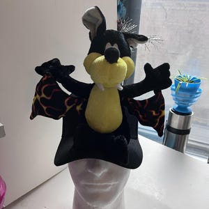 May include: A plush bat hat with a black body, yellow belly, and large white teeth. The bat's wings are black with a red and yellow flame pattern. The hat is displayed on a white mannequin head.