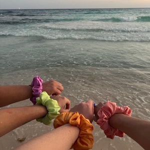 May include: A group of arms wearing colorful scrunchies on a beach. The scrunchies are in shades of pink, green, orange, and peach. The ocean waves are in the background.
