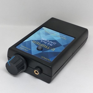 May include: Black electronic device labeled "E-TRIFIER Bob Beck ZAPPER" with a blue and white geometric design. Features a knob labeled "ON/OFF INTENSITY" and an output jack. The device is rectangular with rounded edges.