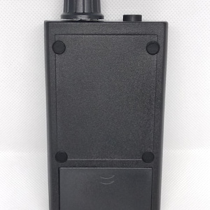 May include: A black electronic device with a textured surface. It features a ridged knob on top, a small button, and a rectangular battery compartment at the bottom. Four small, round rubber feet are visible.