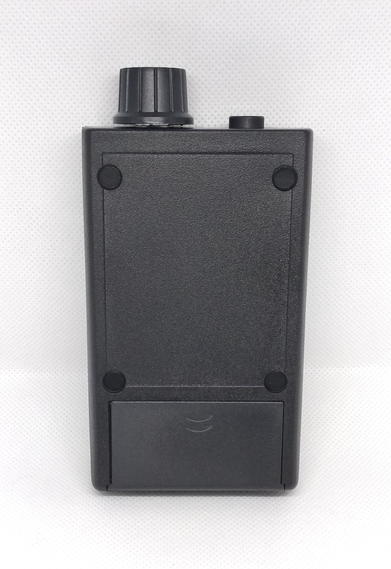 May include: A black electronic device with a textured surface. It features a ridged knob on top, a small button, and a rectangular battery compartment at the bottom. Four small, round rubber feet are visible.